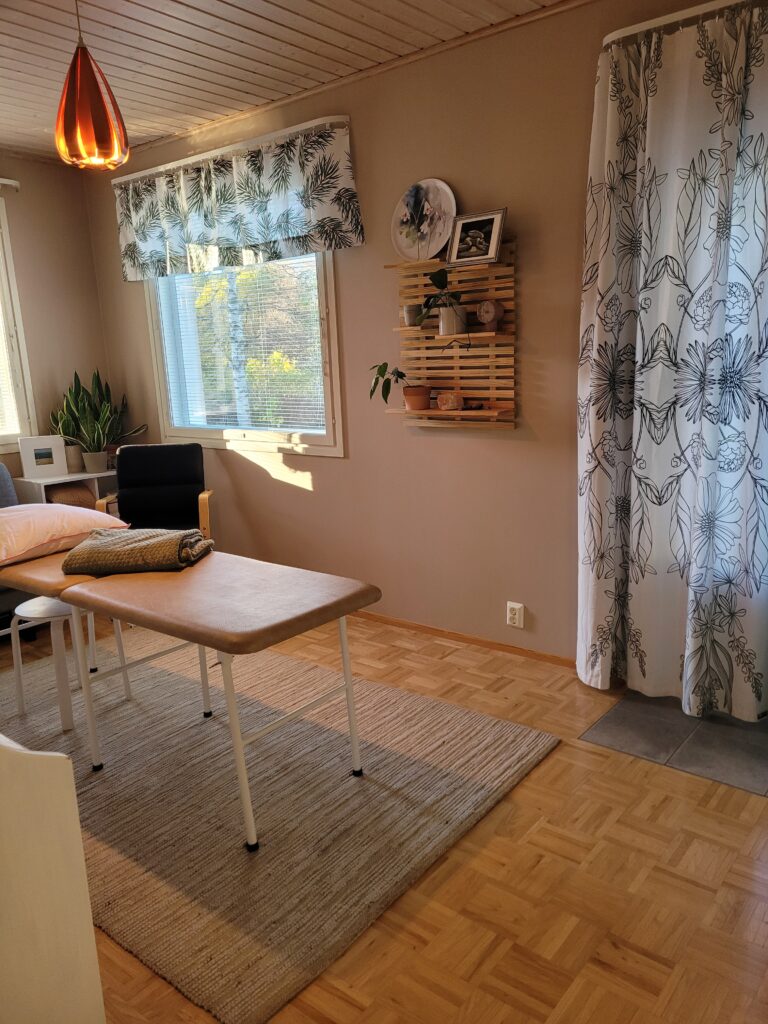 Hoitohuone, keskellä hoitopöytä, jonka päällä on tyyny ja viltti. Huoneessa näkyy kaksi ikkunaa ja ovi.