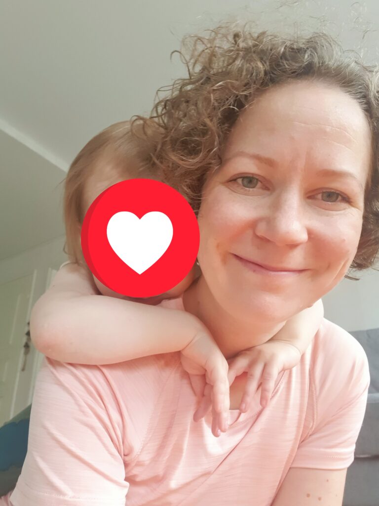 Susanna Slesar jumpalla lapsi selässään. Lapsen kasvot peitetty sydän-ikonilla.